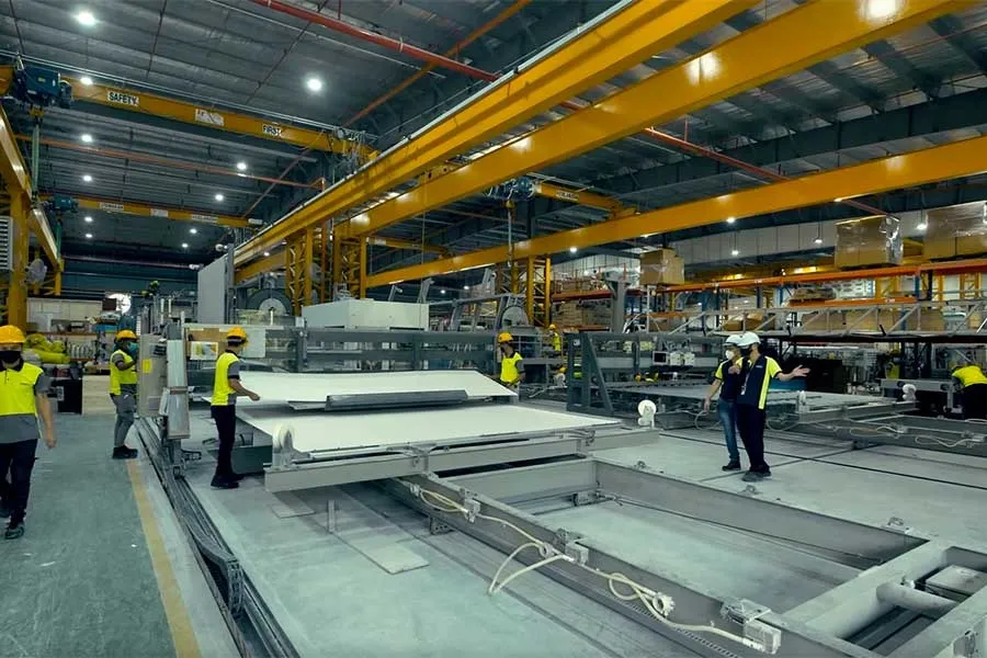 A factory floor with overhead gantry cranes, where workers in hi-vis move a large flat panel along a steel casting bed.